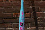 A light blue baseball bat with pink and black graphics, standing vertically against a red-brown brick wall background.