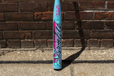 A light blue softball bat with a colorful graffiti-style graphic and 'WORTH' branding, standing upright on a concrete ground against a brick wall background.