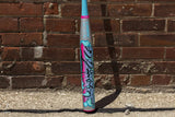 A light blue and gray baseball bat with vibrant graffiti-style pink, purple, and black lettering, standing upright against a red-brown brick wall on a concrete ground.