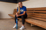 A woman sitting on a wooden bench in a sports locker room, wearing a blue 'DARK' athletic t-shirt, black leggings, and white sneakers while holding a blue and black baseball bat.