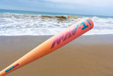 A pink and orange baseball bat with 'NAV-1' and 'PASTIN' branding, resting on a sandy beach with ocean waves in the background.