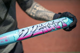 A close-up of a light blue baseball bat with a pink and black graffiti-style graphic, held by a person wearing a black glove on an outdoor field.