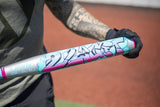 A close-up of a person holding a silver Legit slo-pitch bat with colorful graffiti-style graphics in pink, blue, and black, wearing a black glove on an outdoor baseball field.