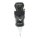 A single Bauer Supreme ice skate boot shown from the back, featuring a black and dark gray carbon-fiber-textured design with white and yellow accents, and a white metal spike at the bottom, set against a plain white background.