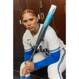 A female baseball player in a white UCLA jersey and blue sleeves holds a Nike U15 UGL baseball bat in a locker room setting.