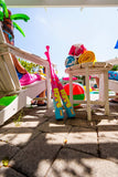 A pair of bright pink and blue plastic pool toys with yellow lettering, leaning against a white lounge chair on a stone patio. A person in a pink swimsuit sits in the chair, and a small table holds rolled towels and a beach ball in a sunny outdoor poolside setting.