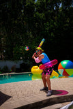 A person standing by a swimming pool wearing a blue sleeveless top, pink shorts, and a yellow cap, holding a colorful inflatable bat and aiming at a floating ball. A pink inflatable ring is around the waist, with pool floats and a large beach ball in the background.